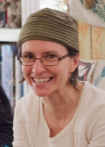 Portrait of Lori Richloff, a white woman with a knit hat and glasses, smiling with photos and paintings in the background